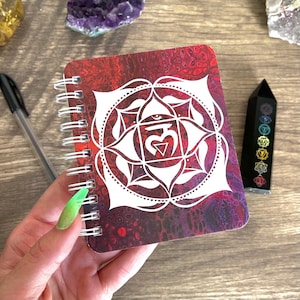 May include: A small red and purple spiral bound notebook with a white mandala design featuring the symbol for the heart chakra, "Anahata".