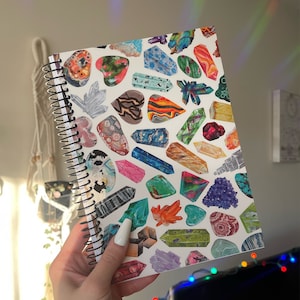 May include: A spiral bound notebook with a white cover featuring a colorful pattern of various crystals and gemstones. The crystals are drawn in a realistic style and include amethyst, rose quartz, and citrine.