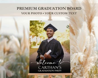 May include: A graduation party welcome sign with a photograph of a graduate in cap and gown, holding a diploma. The sign includes the text "Welcome to Carthan's Graduation Party" and "Premium Graduation Board". The sign is displayed on a wooden easel.