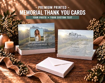 May include: A stack of memorial thank you cards featuring a mountain landscape at sunset, a photo frame, and the text DANA LYNNE FORTESCUE. The cards include a thank you message and are accompanied by a stack of plain white envelopes on a wooden surface.