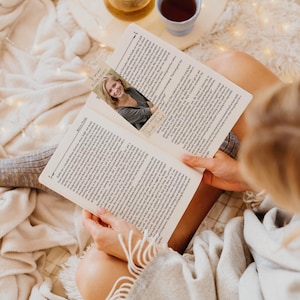 Pode incluir: Uma pessoa lendo um livro com um marcador de p&aacute;gina com uma mulher sorrindo. O livro est&aacute; aberto, revelando texto impresso. Uma x&iacute;cara de ch&aacute; e uma pequena bandeja est&atilde;o no fundo, criando uma cena de leitura aconchegante.