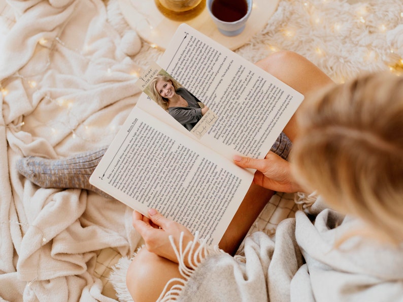 Pode incluir: Uma pessoa lendo um livro com um marcador de p&aacute;gina com uma mulher sorrindo. O livro est&aacute; aberto, revelando texto impresso. Uma x&iacute;cara de ch&aacute; e uma pequena bandeja est&atilde;o no fundo, criando uma cena de leitura aconchegante.