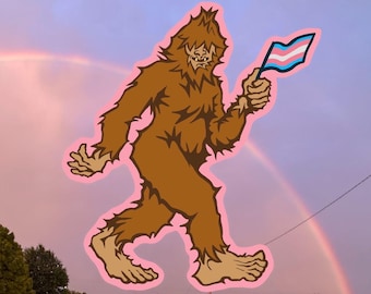 Könnte beinhalten: Eine braune Bigfoot-Cartoon-Illustration mit rosa Umrandung, die eine Transgender-Pride-Flagge hält. Das Bild ist vor dem Hintergrund eines Regenbogens und eines rosa-violetten Himmels platziert.