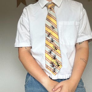 May include: A short-sleeved white button-down shirt paired with a necktie featuring diagonal yellow, white, and grey stripes. The tie is decorated with a series of embroidered black ants scattered across the fabric in a downward pattern.