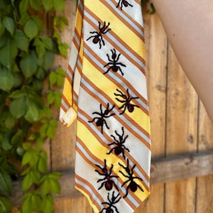 May include: A necktie featuring a diagonal striped pattern in yellow, white, and orange. The tie is decorated with multiple embroidered black ant motifs scattered across the surface, each featuring a small red accent on the abdomen area.