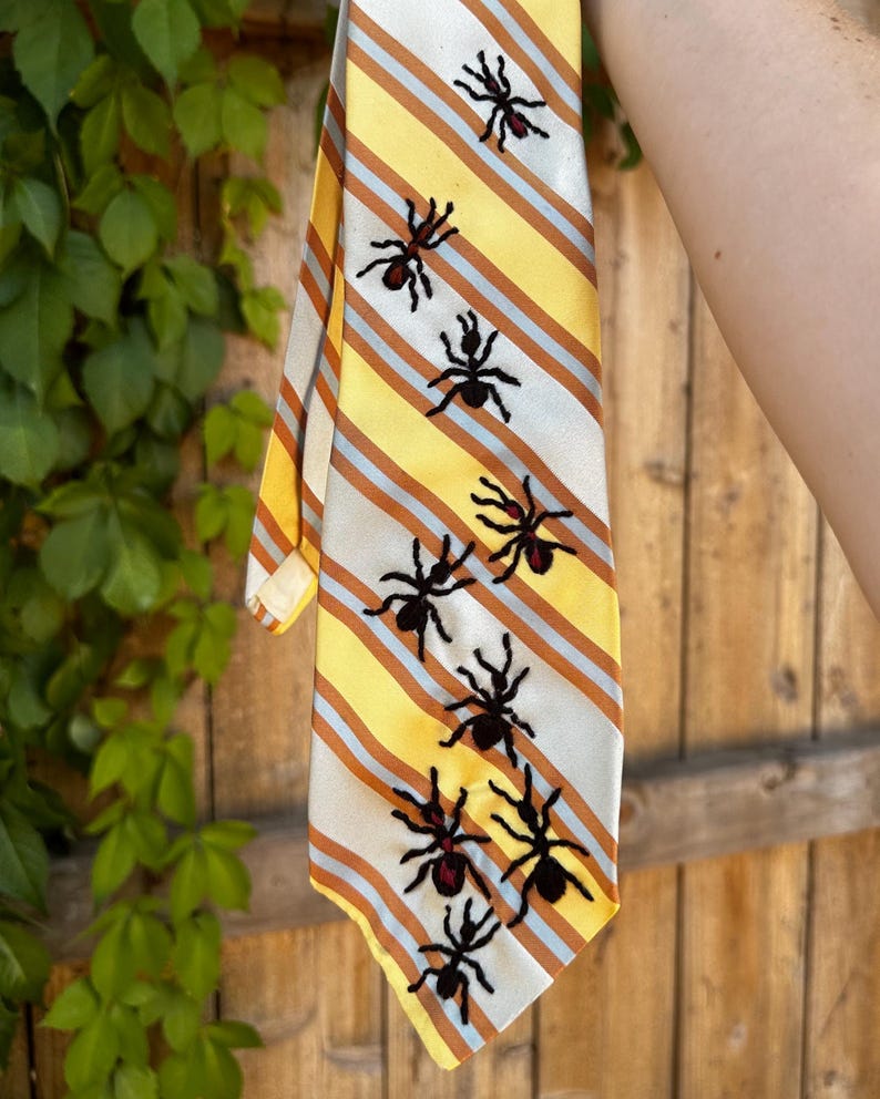 May include: A necktie featuring a diagonal striped pattern in yellow, white, and orange. The tie is decorated with multiple embroidered black ant motifs scattered across the surface, each featuring a small red accent on the abdomen area.