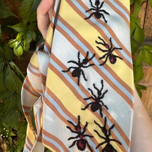 May include: A necktie featuring a diagonal striped pattern in yellow, light blue, and brown. The tie is decorated with embroidered black ant motifs, some of which include small red accents on their bodies, arranged in a scattered pattern.