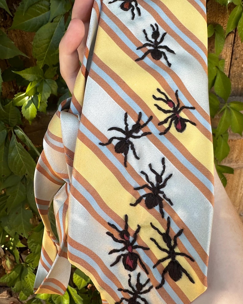 May include: A necktie featuring a diagonal striped pattern in yellow, light blue, and brown. The tie is decorated with embroidered black ant motifs, some of which include small red accents on their bodies, arranged in a scattered pattern.