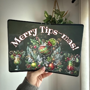 May include: A decorative sign with the text "Merry Tips-mas!" features festive cocktail glasses with holiday decorations. The sign is held up by a person. The background is a dark green color.