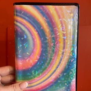 May include: A holographic passport holder with a swirling rainbow pattern and a black border.