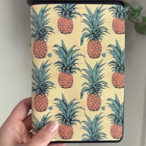 May include: A rectangular notebook cover featuring a repeating pattern of pink pineapples with blue-green leaves on a yellow background. The cover has a dark blue border.