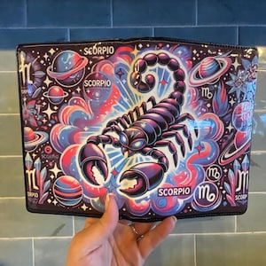 May include: A black wallet featuring a vibrant illustration of a purple scorpion, planets, and the word "SCORPIO". The design incorporates blue, pink, and white accents, creating a cosmic theme. The wallet is held against a blue tile background.