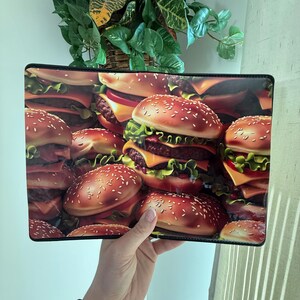 May include: A rectangular clutch featuring a vibrant, detailed print of stacked hamburgers. The design showcases sesame seed buns, cheese, lettuce, and juicy burger patties. The clutch has a black border and is held by a hand.