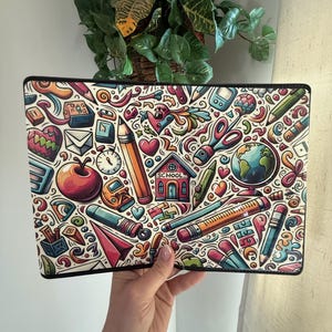May include: A rectangular, white laptop sleeve with a black border, covered in a colorful, cartoon-style school-themed design. The design includes pencils, apples, hearts, a globe, and a schoolhouse. The sleeve is held by a person.