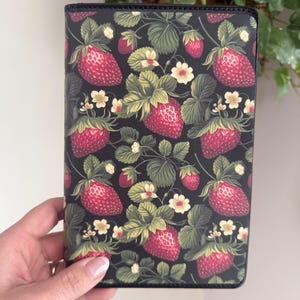 May include: A black journal cover featuring a repeating pattern of ripe red strawberries, green leaves, and small white flowers. The design is set against a dark background, creating a vibrant contrast. The cover is held by a hand.