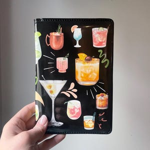 May include: Black leather wallet with a colorful illustration of various cocktails and drinks. The wallet has a stitched edge and a flap closure.