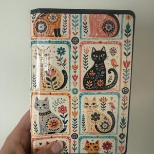 May include: A rectangular notebook cover featuring a grid pattern of illustrated cats in various colors, including black, cream, and orange. Each cat is decorated with floral and geometric patterns, surrounded by small birds and botanical motifs.