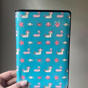 May include: A black passport holder with a turquoise background and a repeating pattern of pink flamingos, white unicorns, seashells, starfish, and crabs.
