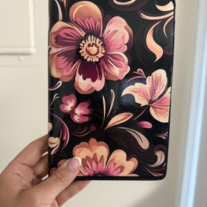 May include: A rectangular passport holder featuring a glossy black background adorned with a vibrant floral pattern. The design includes large, stylized flowers in shades of peach, soft pink, and deep magenta, accented by flowing, dark-toned leaf motifs.