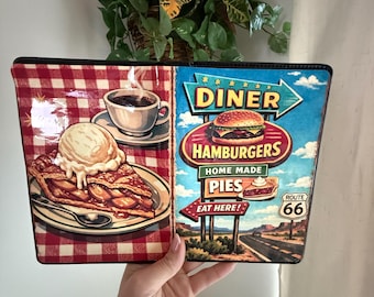 Può includere: Un cartello rettangolare a tema diner con una fetta di torta di mele, caffè e un cartello della Route 66. Il cartello ha un motivo a scacchi rossi e bianchi sul lato sinistro e una scena di diner sul lato destro.