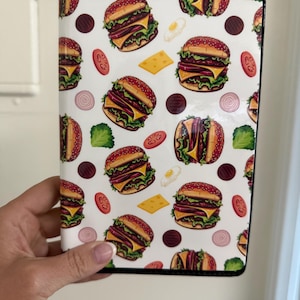 May include: A rectangular passport holder featuring a repeating pattern of cheeseburgers, tomato slices, lettuce leaves, onion rings, cheese squares, and fried eggs on a white background with a black border trim along the edges.