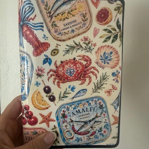 May include: A rectangular notebook cover featuring a cream background with a colorful pattern of seafood, including a red crab, a lobster, and tins of sardines labeled SARDINE LIMONE & BASILICO and AMALFI SARDINE PICCANTI, alongside lemons, olives, and shells.