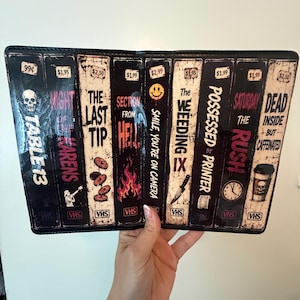 May include: A rectangular black wallet featuring a graphic print of nine vintage VHS tape spines. The titles include Table 13, Karens, The Last Tip, Section From Hell, Smile You're On Camera, The Weeding, Possessed By Printer, Rush, and Dead But Caffeinated.