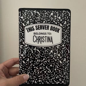 May include: A black and white server book with a composition notebook design. The text on the cover reads "THIS SERVER BOOK BELONGS TO: CHRISTINA". The book is held by a hand.