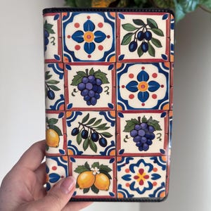 May include: A decorative book cover featuring a colorful tile design. The pattern includes images of grapes, lemons, and olives, set against a white background with blue, orange, and red accents. The cover has a black border.