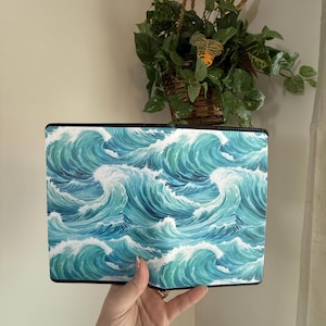May include: A rectangular notebook cover featuring a watercolor design of ocean waves in shades of blue and teal. The waves are depicted with white foam, creating a dynamic and artistic pattern. The cover has a black border.