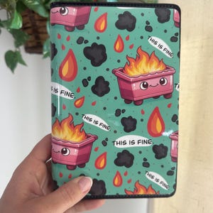 May include: A teal notebook cover with a repeating pattern of a pink trash can on fire, flames, and black smoke. The text "THIS IS FINE" is written on the cover.