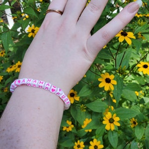 Puede incluir: Un brazalete de cuentas blancas con letras rosas que dice "I ♥ HAHAHA FUCK". El brazalete se lleva en la muñeca de una persona sobre un fondo de flores amarillas.