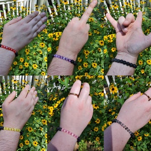 Puede incluir: Seis pulseras de roca de lava diferentes con cuentas y letras de varios colores. Las pulseras se llevan en las muñecas de seis personas diferentes. Las personas están de pie frente a un fondo de flores amarillas.