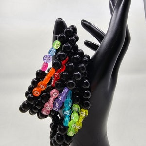 May include: A set of colorful beaded bracelets with black beads and white lettering. The bracelets are stacked on top of each other and are held in a black hand.