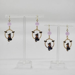 Puede incluir: Un par de pendientes de tono dorado con una cuenta morada y un dije de una bruja en un círculo con murciélagos.