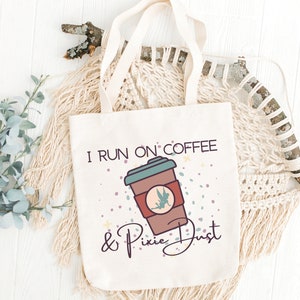 May include: A white tote bag with the text "I run on coffee & pixie dust" printed on it. The text is in brown and black lettering. A cartoon image of a coffee cup with a fairy inside is above the text.