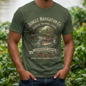 Peut inclure: T-shirt vert olive avec un graphisme de style vintage. Le graphisme affiche "Jungle Navigation Co. Skipper Training School" et comprend un bateau, des éléphants et les mots "Experienced Skipper". Le t-shirt est porté par une personne devant une rivière.