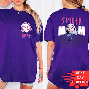May include: A purple t-shirt featuring a Spider-Gwen design. The front displays a white and pink Spider-Gwen mask with the word "NAME" below. The back has "SPIDER MOM" above a Spider-Gwen graphic. The shirt includes a "NEXT DAY SHIPPING" graphic.