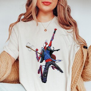 May include: A white t-shirt with a graphic of a red and blue Spider-Man character playing a guitar. The character is wearing a blue jacket and black pants.