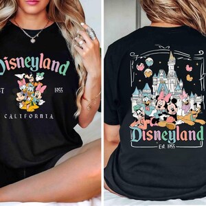 Puede incluir: Camiseta negra con un gr&aacute;fico de Disneyland colorido en la parte delantera y trasera. El gr&aacute;fico delantero presenta a Mickey Mouse y sus amigos, con el texto "Disneyland EST 1955 CALIFORNIA". El gr&aacute;fico trasero presenta a Mickey Mouse y sus amigos frente al castillo de Disneyland, con el texto "Disneyland - Est. 1955 - ".