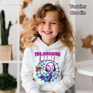 Op de afbeelding: Witte hoodie voor peuters met een superheldenafbeelding, spinnenweb en de tekst "THE AMAZING NAME". De hoodie heeft een capuchon en lange mouwen. De tekst "Toddler Hoodie" is ook aanwezig.