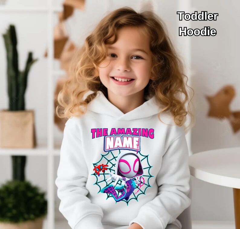Op de afbeelding: Witte hoodie voor peuters met een superheldenafbeelding, spinnenweb en de tekst "THE AMAZING NAME". De hoodie heeft een capuchon en lange mouwen. De tekst "Toddler Hoodie" is ook aanwezig.