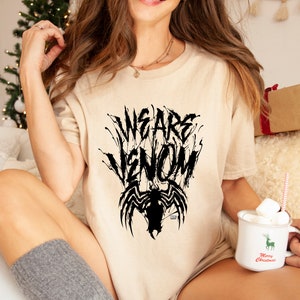 May include: Beige t-shirt with the text "WE ARE VENOM" in a black, stylized font, with a black Venom graphic below. The shirt is a casual style, suitable for everyday wear.