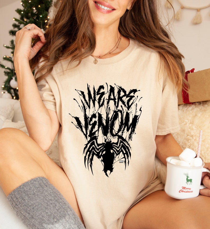 May include: Beige t-shirt with the text "WE ARE VENOM" in a black, stylized font, with a black Venom graphic below. The shirt is a casual style, suitable for everyday wear.