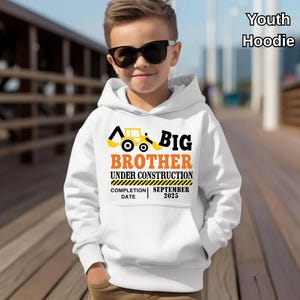 May include: White youth hoodie with a front pocket, featuring a construction-themed design. The design includes a yellow excavator, the words "BIG BROTHER UNDER CONSTRUCTION," and the completion date "SEPTEMBER 2025."