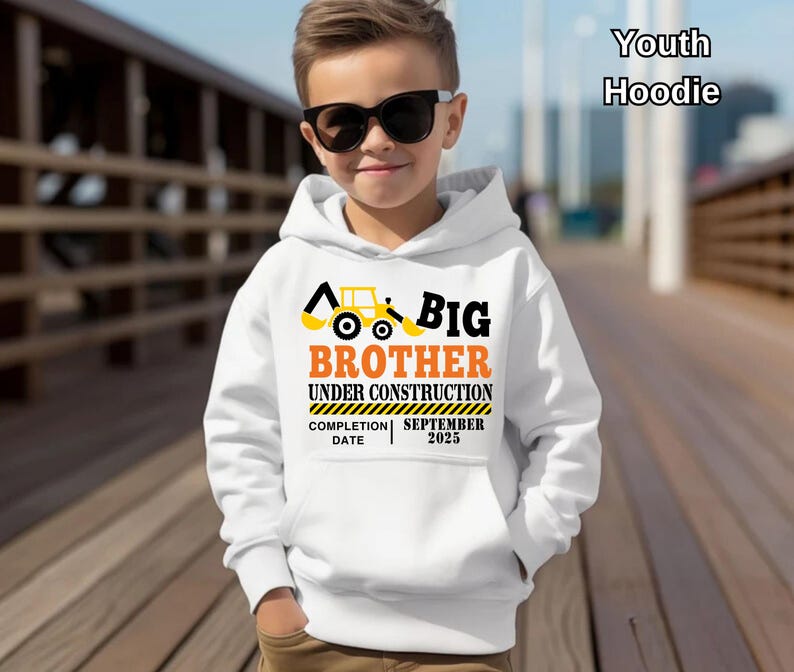 May include: White youth hoodie with a front pocket, featuring a construction-themed design. The design includes a yellow excavator, the words "BIG BROTHER UNDER CONSTRUCTION," and the completion date "SEPTEMBER 2025."