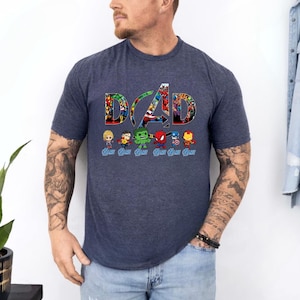Peut inclure: Un t-shirt &agrave; manches courtes bleu chin&eacute; fonc&eacute; avec le mot DAD en grandes lettres remplies d'images de super-h&eacute;ros. Sous le texte se trouvent six petites figurines de super-h&eacute;ros de style chibi, chacune accompagn&eacute;e du mot DAD &eacute;crit en bleu.