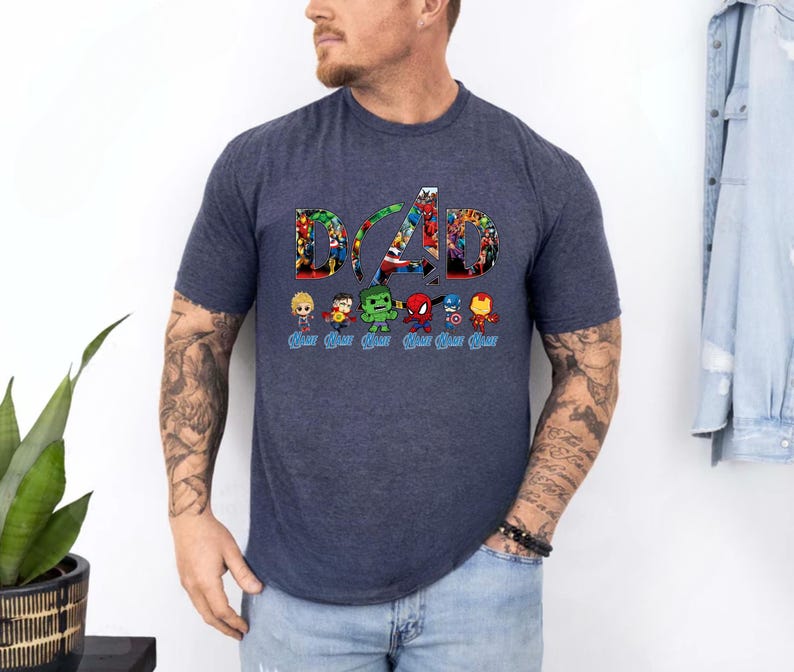 Peut inclure: Un t-shirt &agrave; manches courtes bleu chin&eacute; fonc&eacute; avec le mot DAD en grandes lettres remplies d'images de super-h&eacute;ros. Sous le texte se trouvent six petites figurines de super-h&eacute;ros de style chibi, chacune accompagn&eacute;e du mot DAD &eacute;crit en bleu.