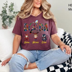 Peut inclure: Un t-shirt &agrave; manches courtes couleur bordeaux chin&eacute; avec le texte DADA rempli de motifs de super-h&eacute;ros. Sous le texte se trouvent trois personnages de style chibi avec les pr&eacute;noms Sophia, Michael et Ethan imprim&eacute;s en jaune en dessous.