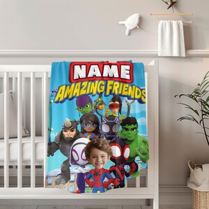 May include: A blue blanket with the text "NAME and his AMAZING FRIENDS" and cartoon superhero characters. The blanket is draped over a white crib. The blanket features a child's face in the center.